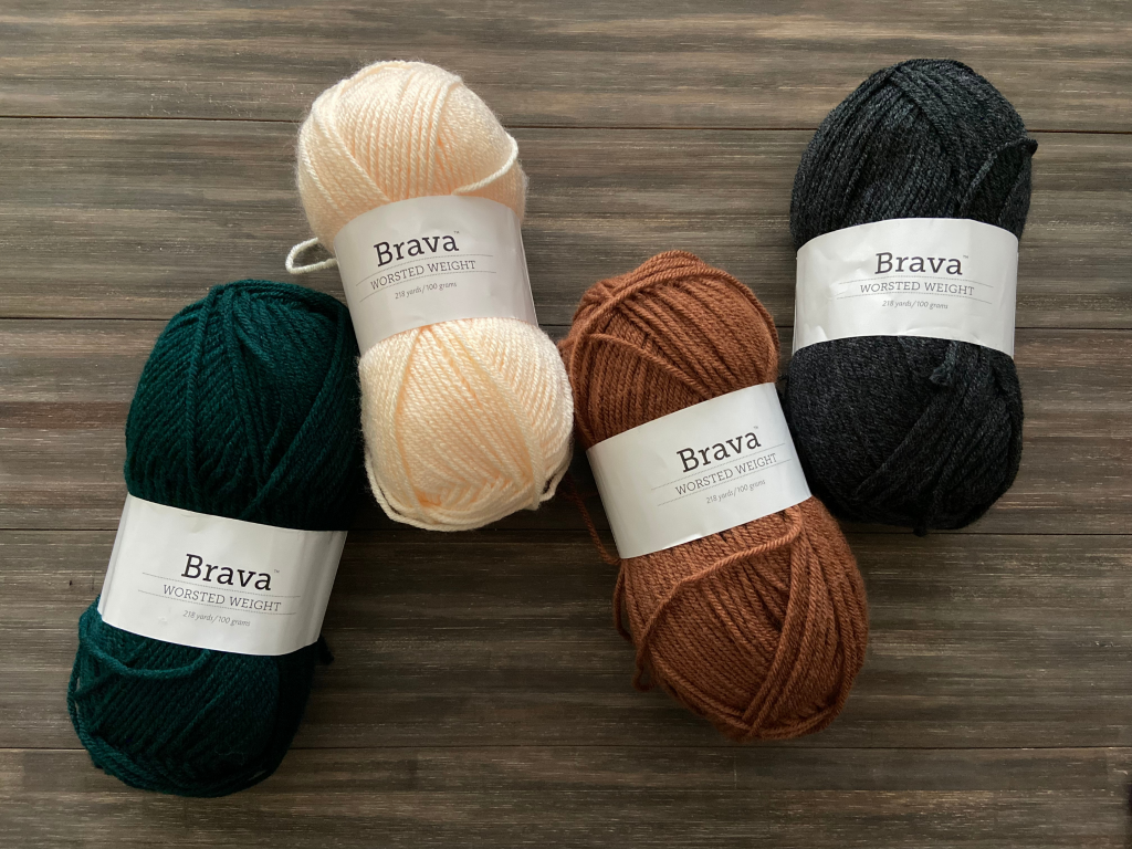 4 skeins of Brava worsted yarn in colors green, cream, brown and charcoal on a wood background