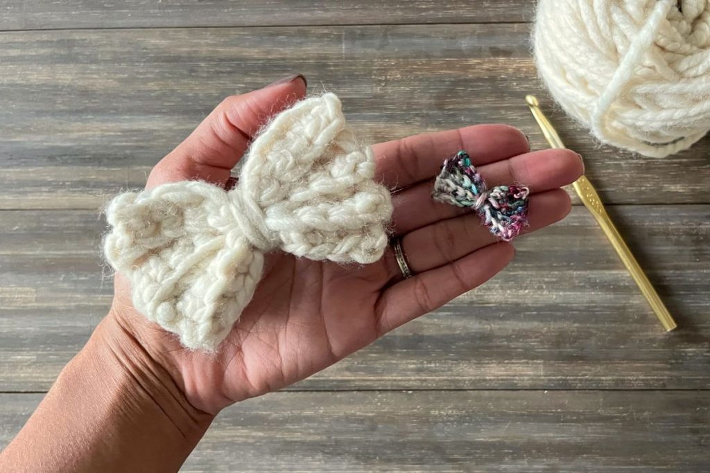hand holding a large cream crochet bow and a small multi color crochet bow in front of wood background