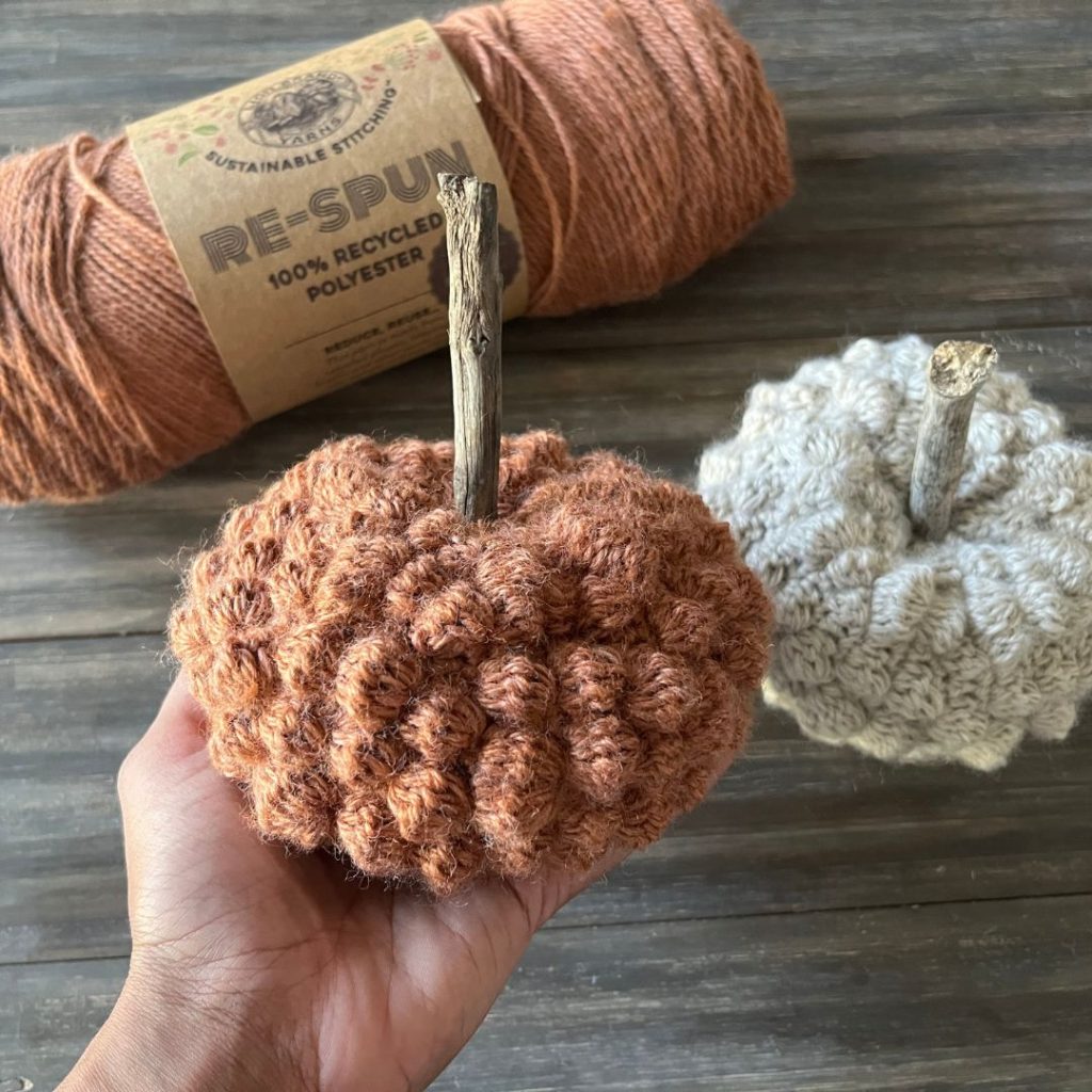 hand holding textured orange crochet pumpkin next to cream crochet pumpkin on wood background