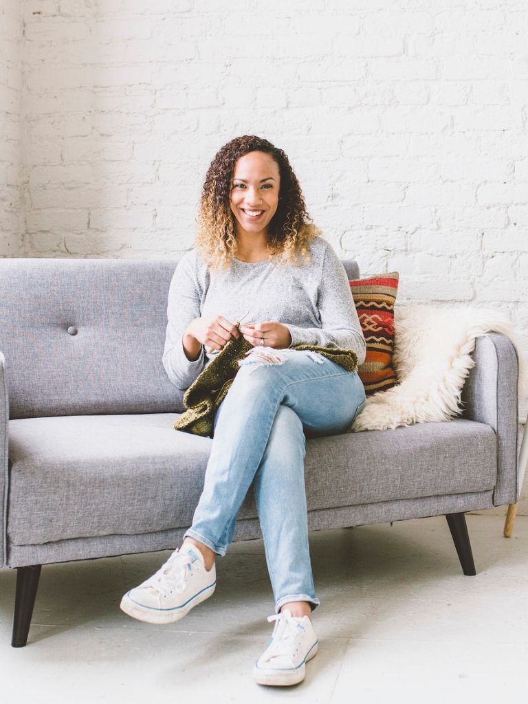 black woman smiling and sitting on gray couch holding green crochet project