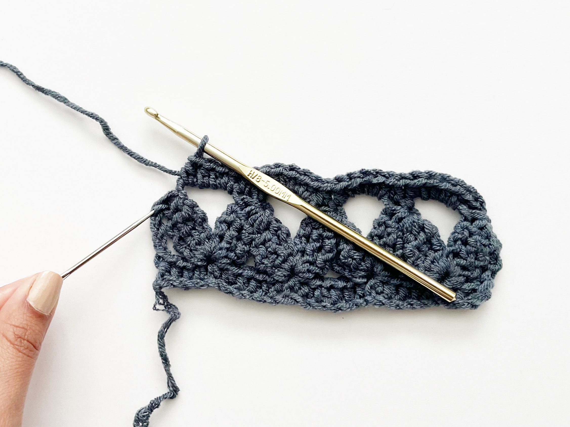 hand using tapestry needle to point to stitch on a blue crochet sample with gold crochet hook 