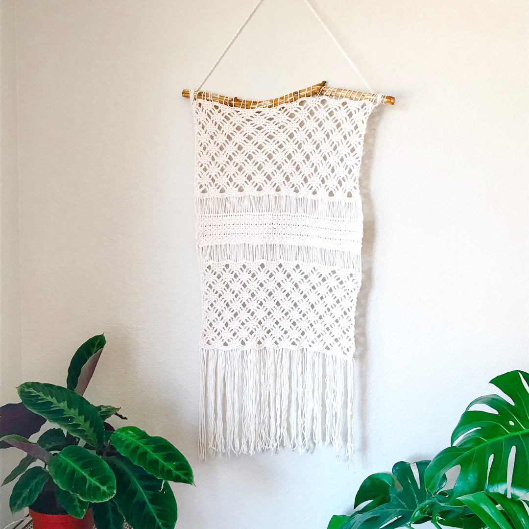 cream crochet wall-hanging on light wood stick near houseplants