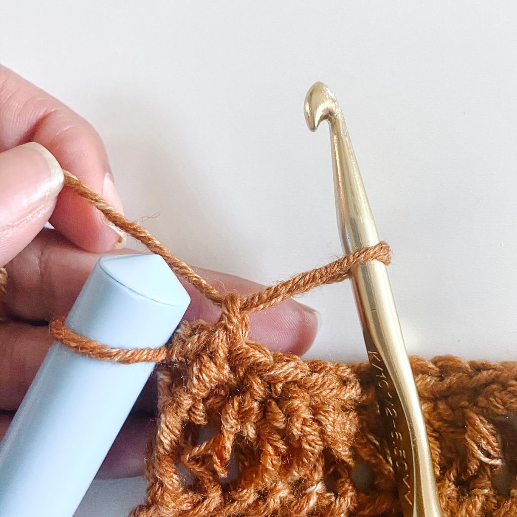 gold crochet hook pulling up long loop in brown yarn with light blue dowel